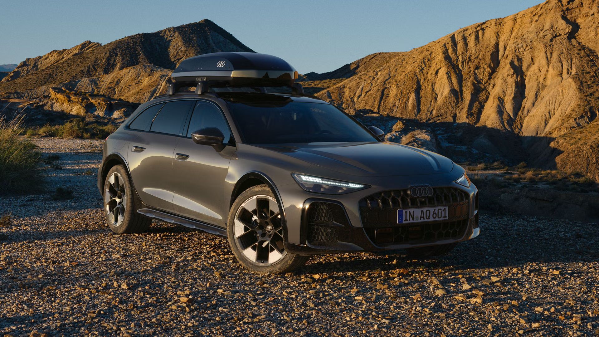 Der Audi A6 allroad schräg von vorn mit brilliantschwarzer Dachbox parkt auf einem Schotterweg. Im Hintergrund eine karge und felsige Landschaft, Sonnenlicht von der Seite taucht die Szenerie in schattige und helle Abschnitte.